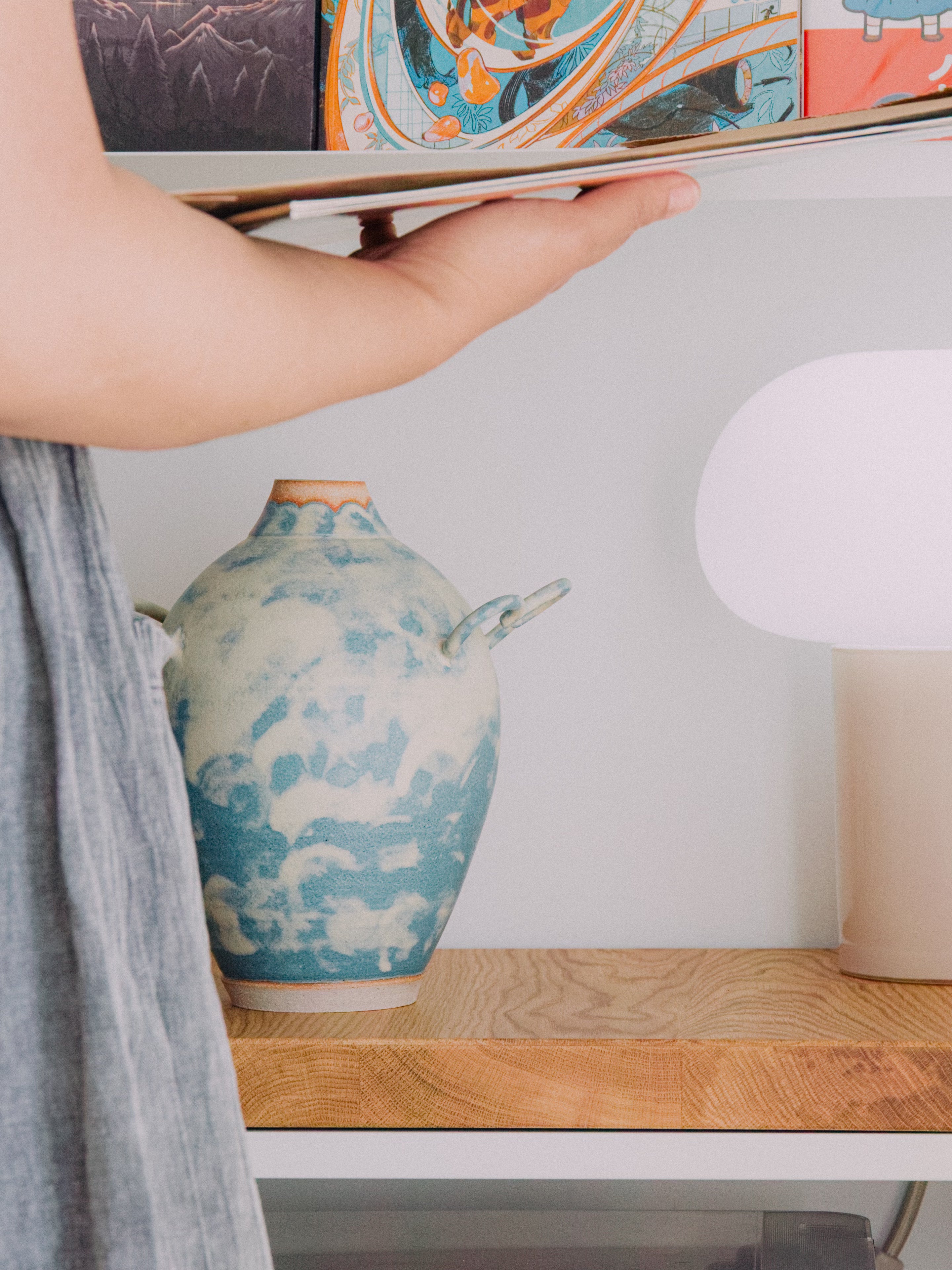 Person holding a tablet above a ceramic vase on a wooden surface with a lamp in the background.